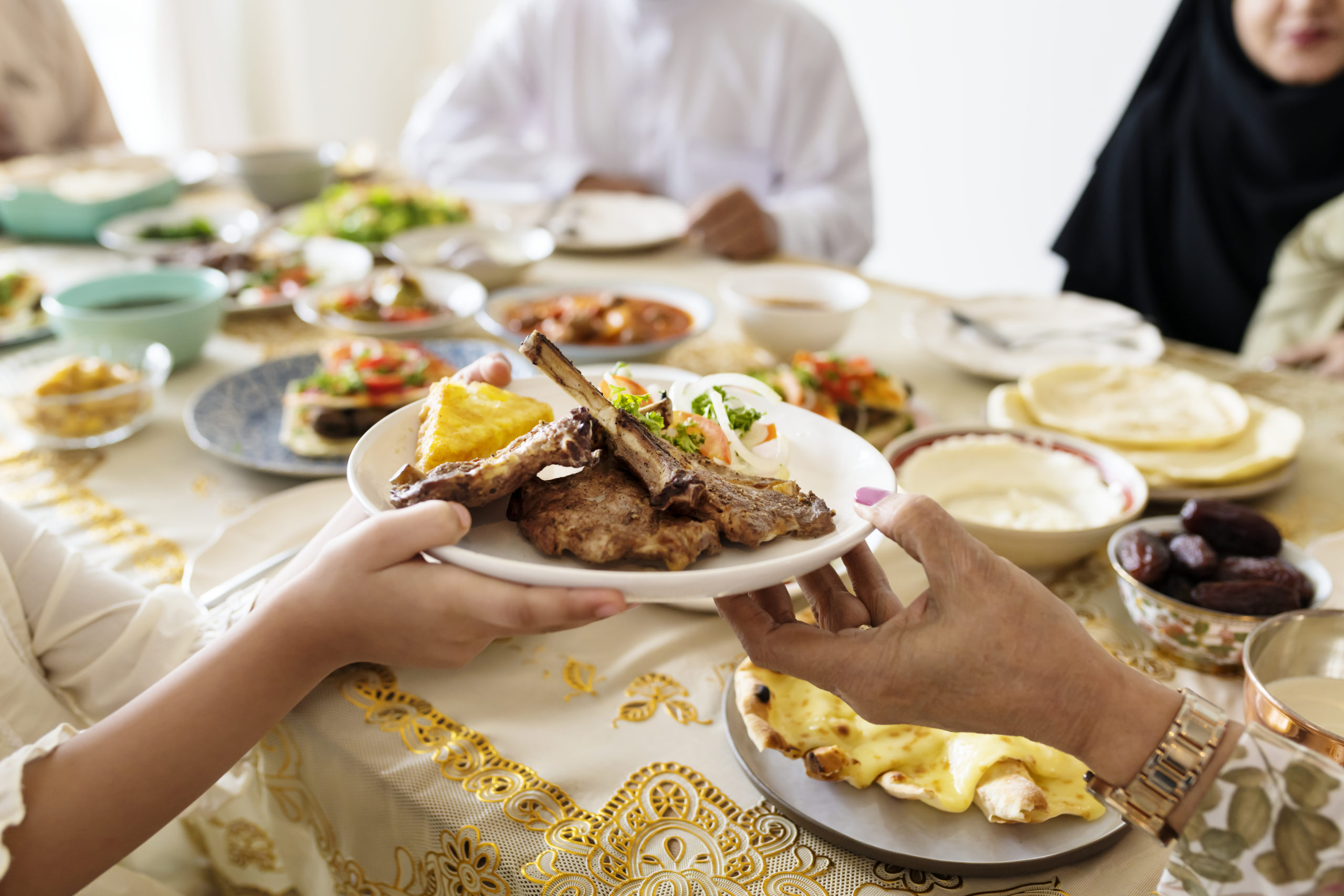 Une famille musulmane partage un repas de l'Aïd autour d'une table. Au premier plan apparaît une main qui tend une assiette contenant des côtelettes d'agneau et de la salade à une autre personne.